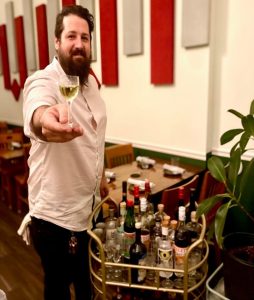 A man in a collared shirt holds a glass of white wine toward the camera. To his right is a bar cart stocked with bottles.