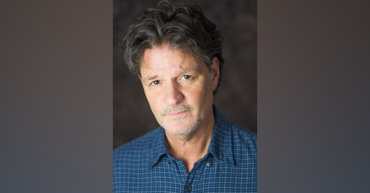 A photo of a white man in a blue collared shirt against a dark background.