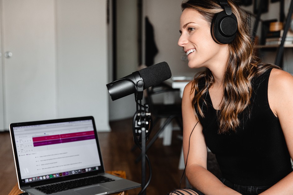 White woman with long hair, wearing black headphones and sitting at a table next to a microphone. A laptop is on the table.