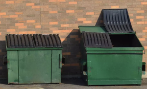 Two green dumpsters with black lids, next to each other by a brick wall. Half the lid is open on the right dumpster.