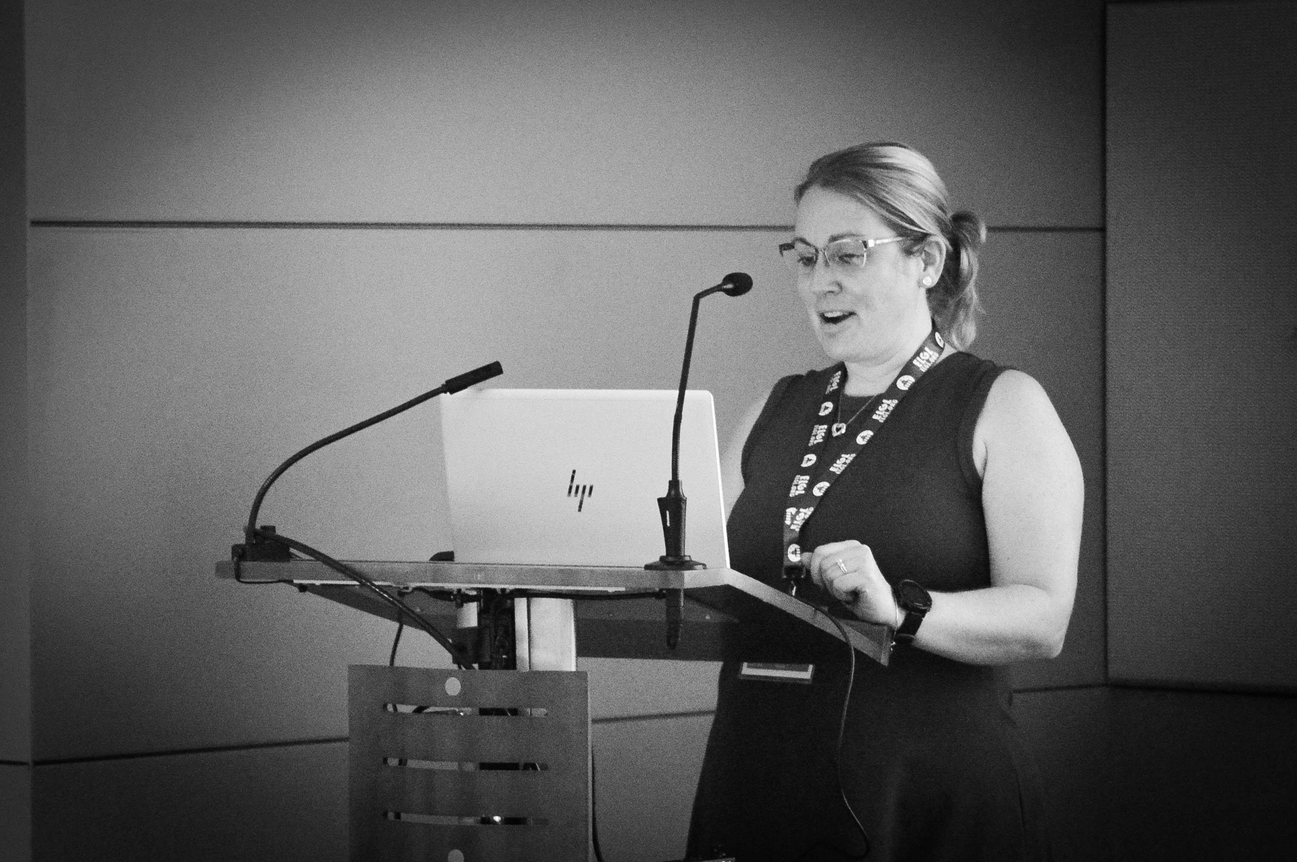 In a black-and-white photo, a woman stands at a lectern presenting during a conference session.