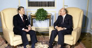 President Ronald Reagan speaks to another dignitary while both are seated in chairs in front of a bookshelf.