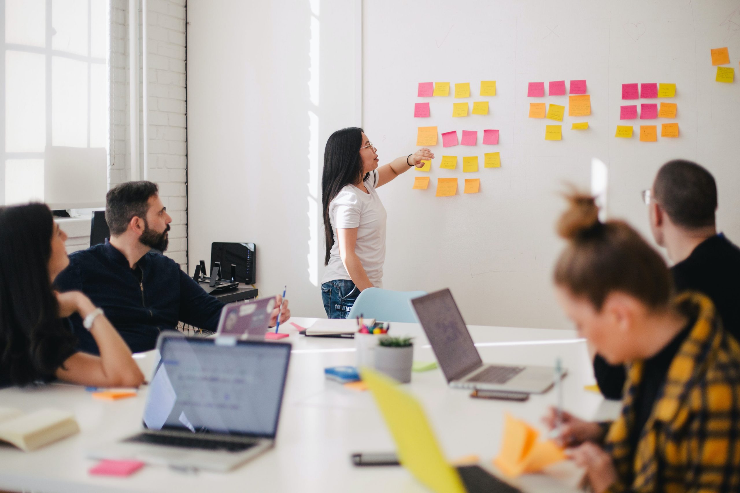 A group of people sit around a table in a conference room. One person is standing at the head of the table pointing to sticky notes displayed on the wall.