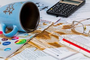 A messy desk with a spilled coffee cup, coffee stains on paper documents, reading glasses, and a calculator.