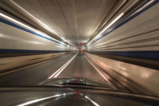 A road tunnel viewed from inside a vehicle, with the hood visible and lights streaking past on the tunnel walls.