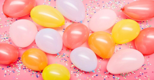 Balloons in pastel colors of pink, yellow, and lilac lie on a pink floor strewn with confetti.