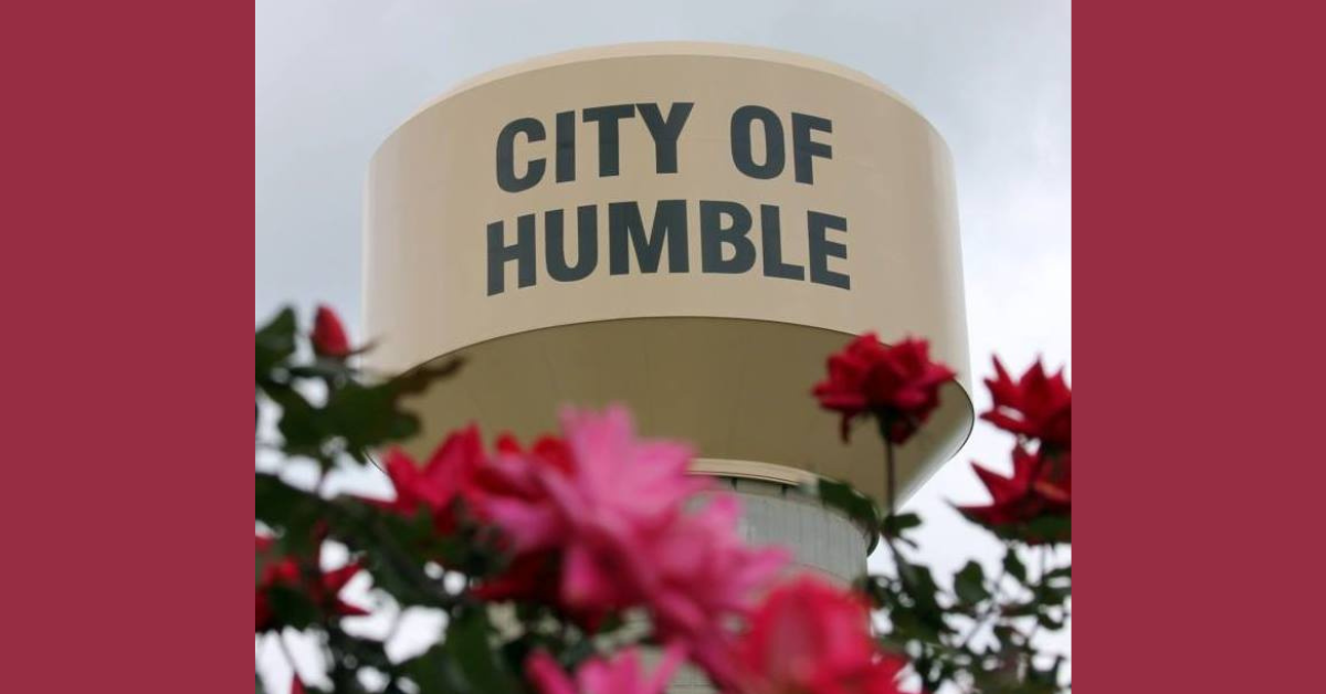 A water tower with the words "City of Humble" painted on the side. In the foreground are magenta flowers.