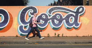 A man walking along a city street in front of a mural that reads "Good" in a looping script font.