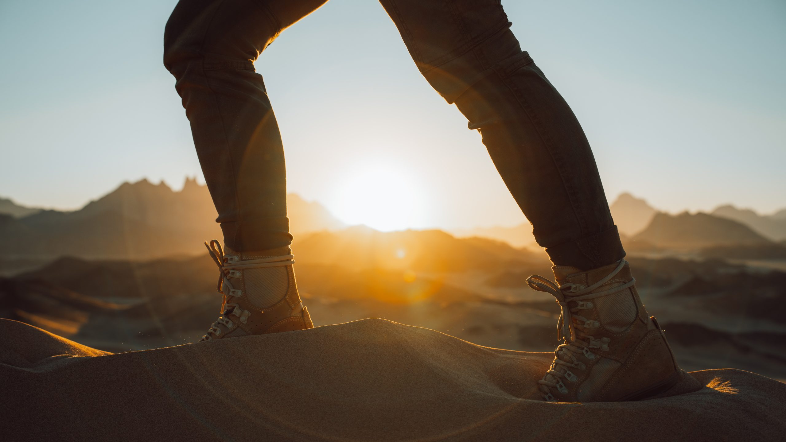 A close-up of a person's legs and hiking boots as they walk through desert sand in Saudi Arabia. In the background is the rising sun.