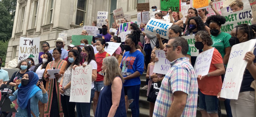 Dozens of people standing on the steps of an institutional or government building, some holding signs with messages about funding and budget.