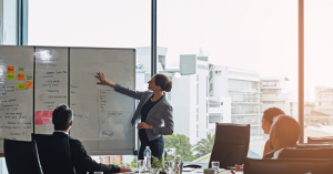 People gathered around a table in a boardroom watch a presenter pointing at a whiteboard.