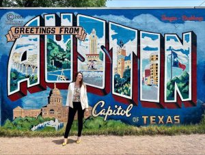 Woman standing in front of colorful "greetings from Austin, capital of Texas" mural.