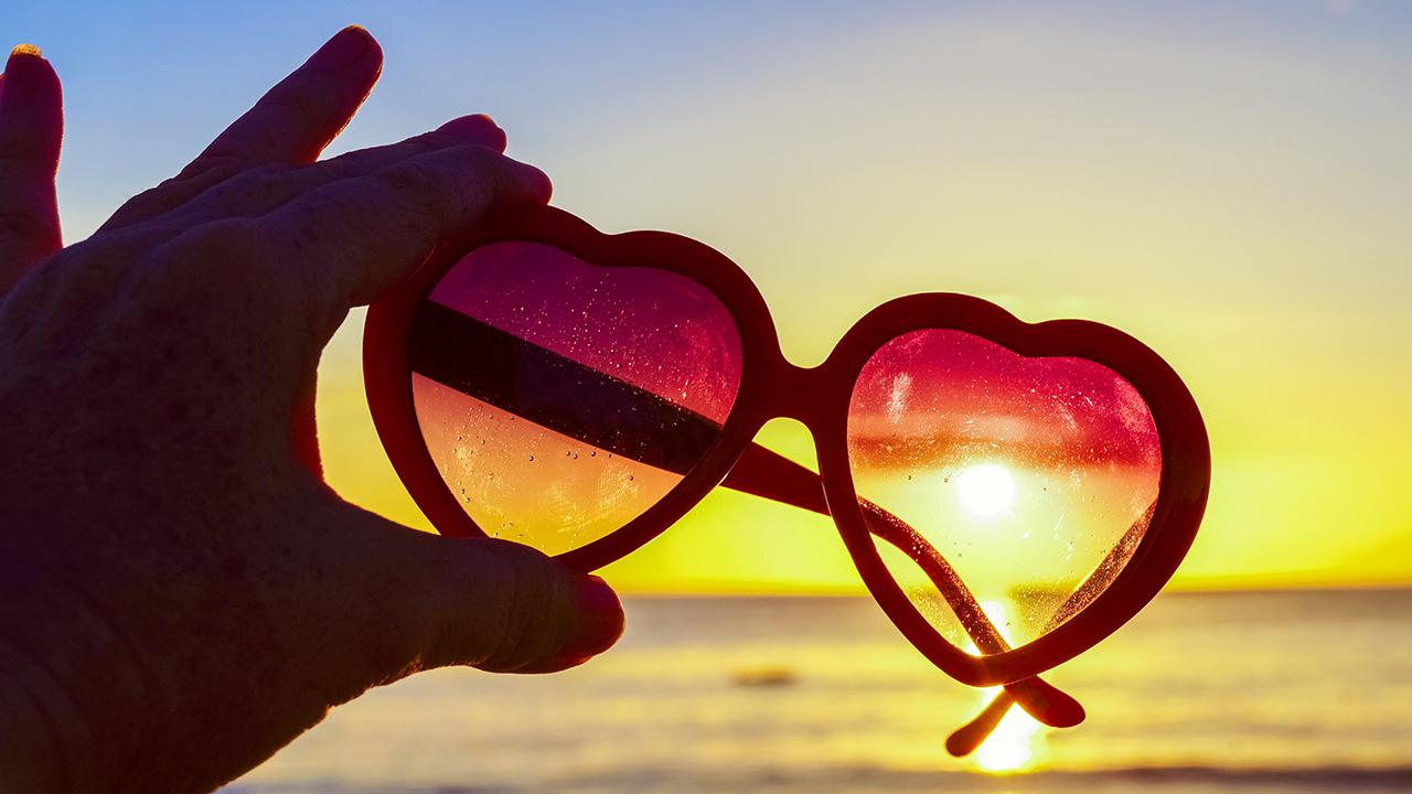 A hand holds a pair of sunglasses with heart-shaped lenses up on a beach to show a sunset through the lenses.