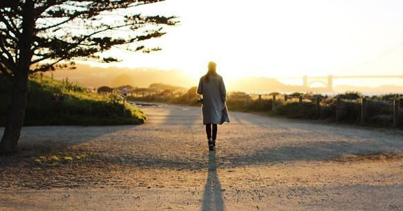 A person walks away from the camera into the light of a sunrise or sunset along an unpaved path. A tree is visible at the left of the image.