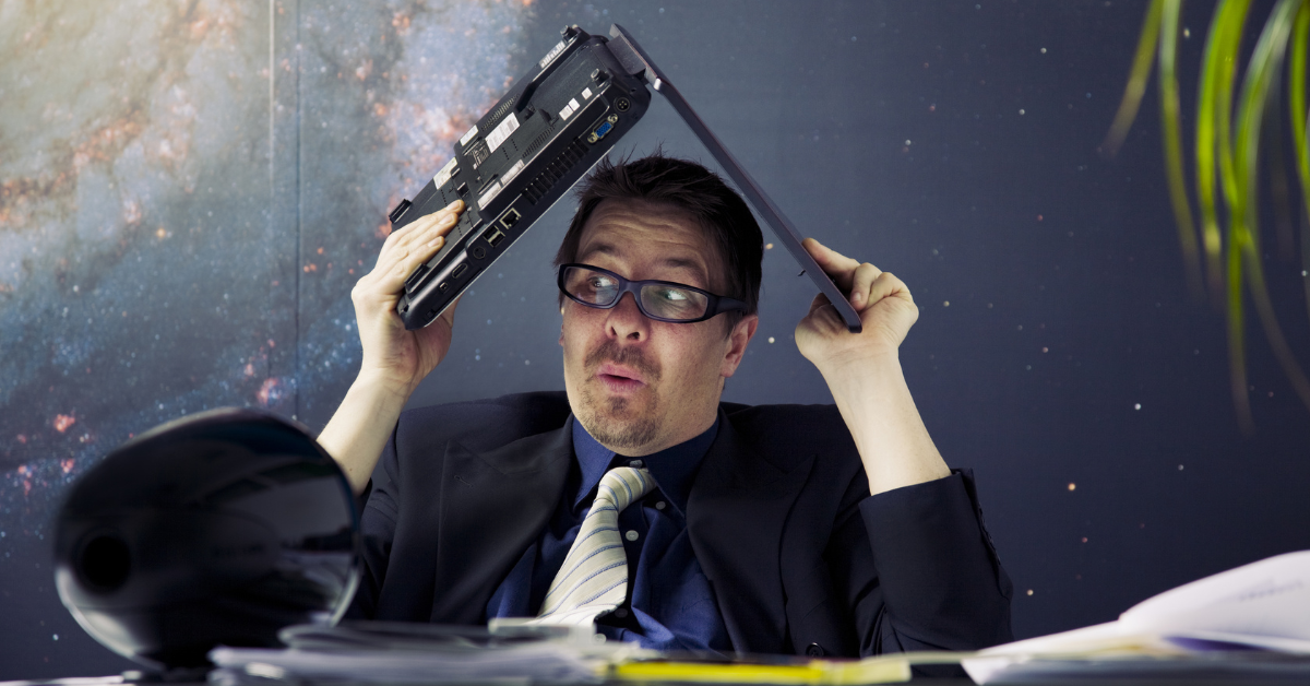 A man wearing a jacket and tie holds laptop over his head. The wall behind him resembles a photo of a galaxy and a palm frond is visible in the right side of the frame.