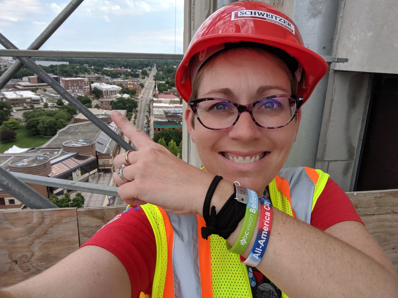 A selfie showing a woman in a hardhat and a safety vest pointing toward a building in the background.