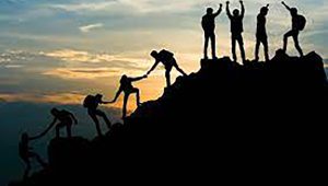 A group of 8 people, shown in silhouette against a rising or setting sun, reach the peak of a mountain.