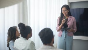 A woman stands at the front of a conference room and speaks to 3 people seated in front of her.