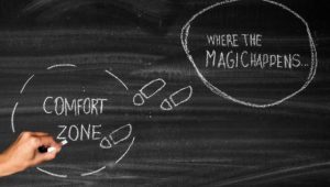 A blackboard with the words "comfort zone" in a word bubble on the left and the words "where the magic happens" in a word bubble on the right. The words are written in chalf and a hand hovers over the words "comfort zone."