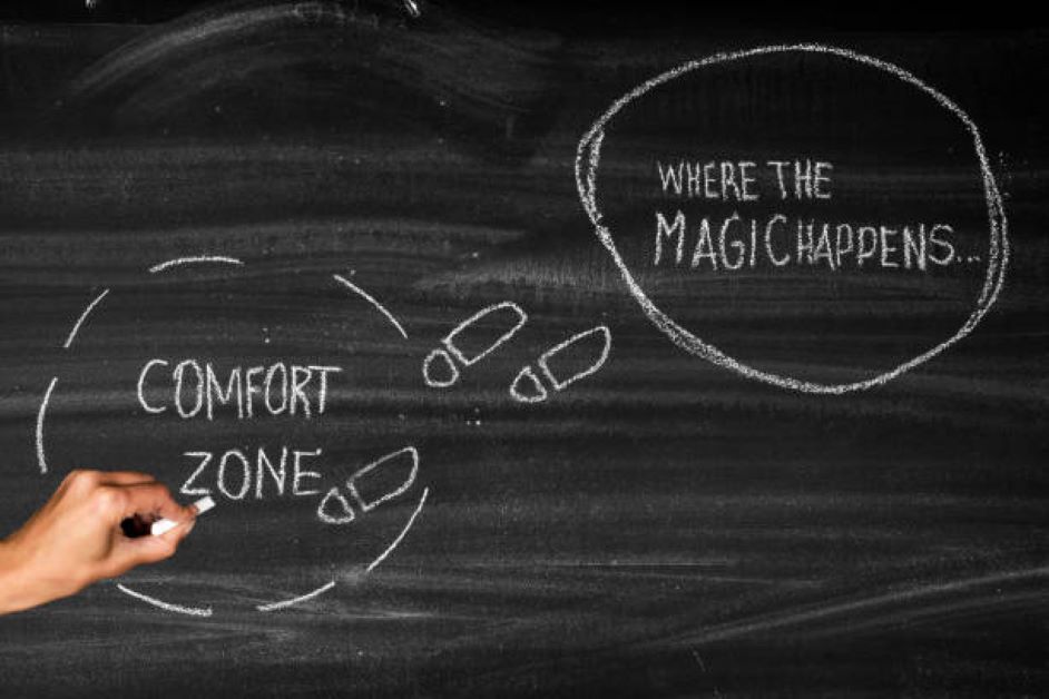 A blackboard with the words "comfort zone" in a word bubble on the left and the words "where the magic happens" in a word bubble on the right. The words are written in chalf and a hand hovers over the words "comfort zone."