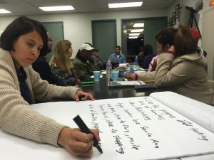 A woman writes on an oversized notepad using a thick black marker. Behind her, other community members talk to each other.