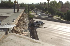 A photo of damage after an earthquake. There is debris from a sunken in bridge and an upside-down car lies below two emergency personnel.