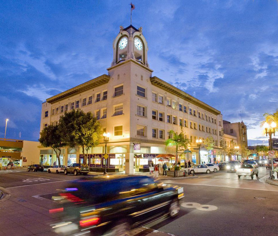 A historic building with a clocktower, the Spurgeon Building in Santa Ana, stands tall as cars zip by on the streets below. The day is turning to night. The sky is deep blue and clouds rush past.