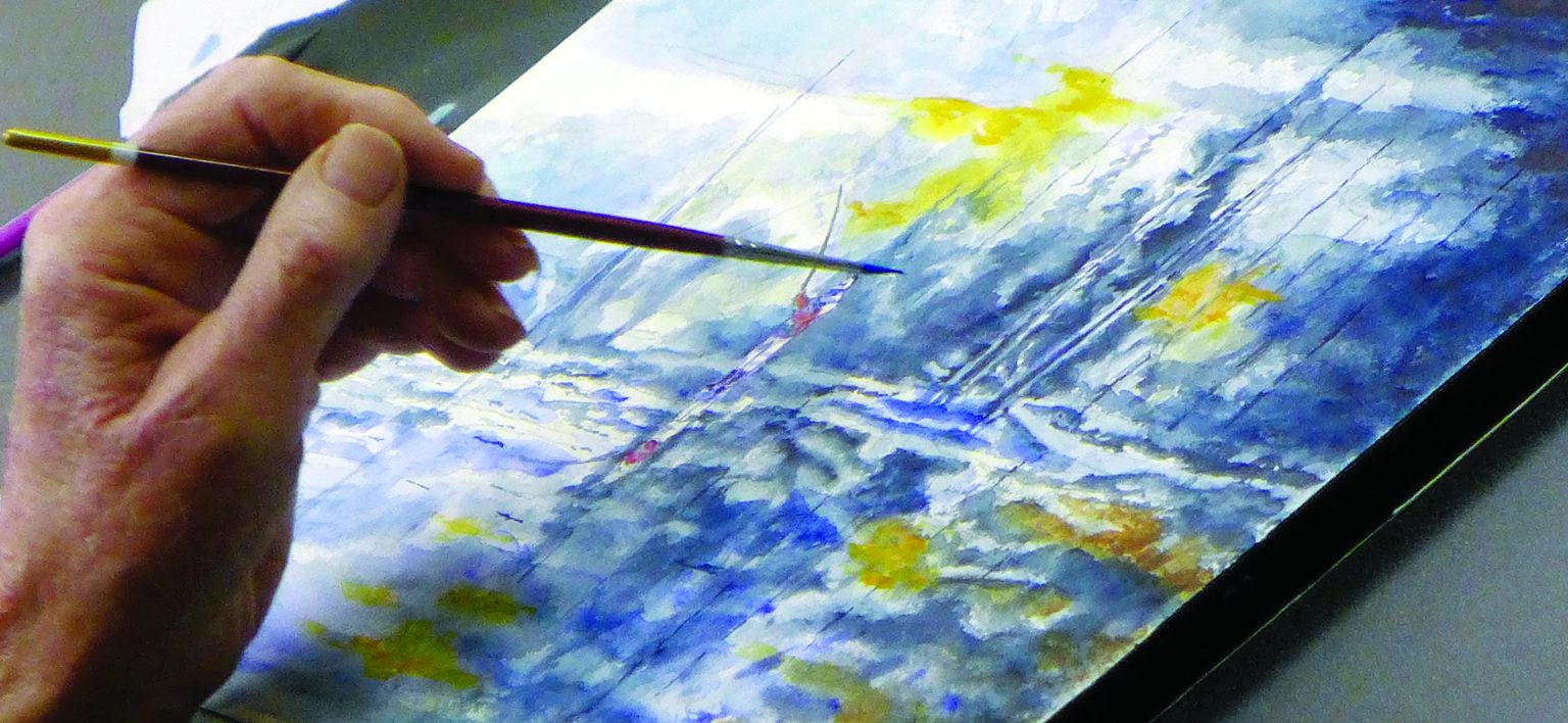 Hand holding a paint brush up to a watercolor painting of a person fishing