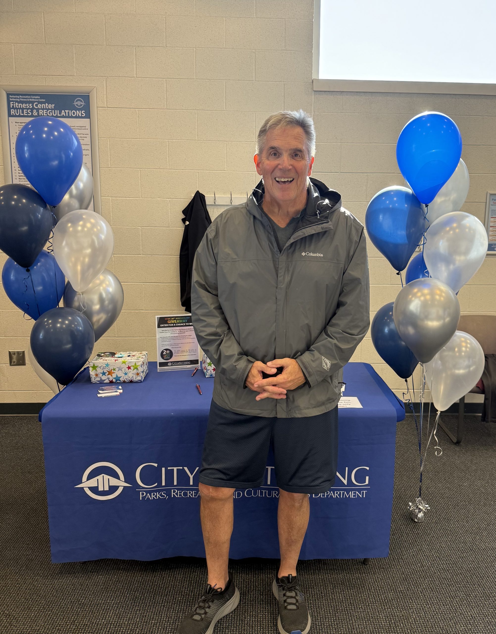 Man in gray jacket stands in front of table with blue and white balloons
