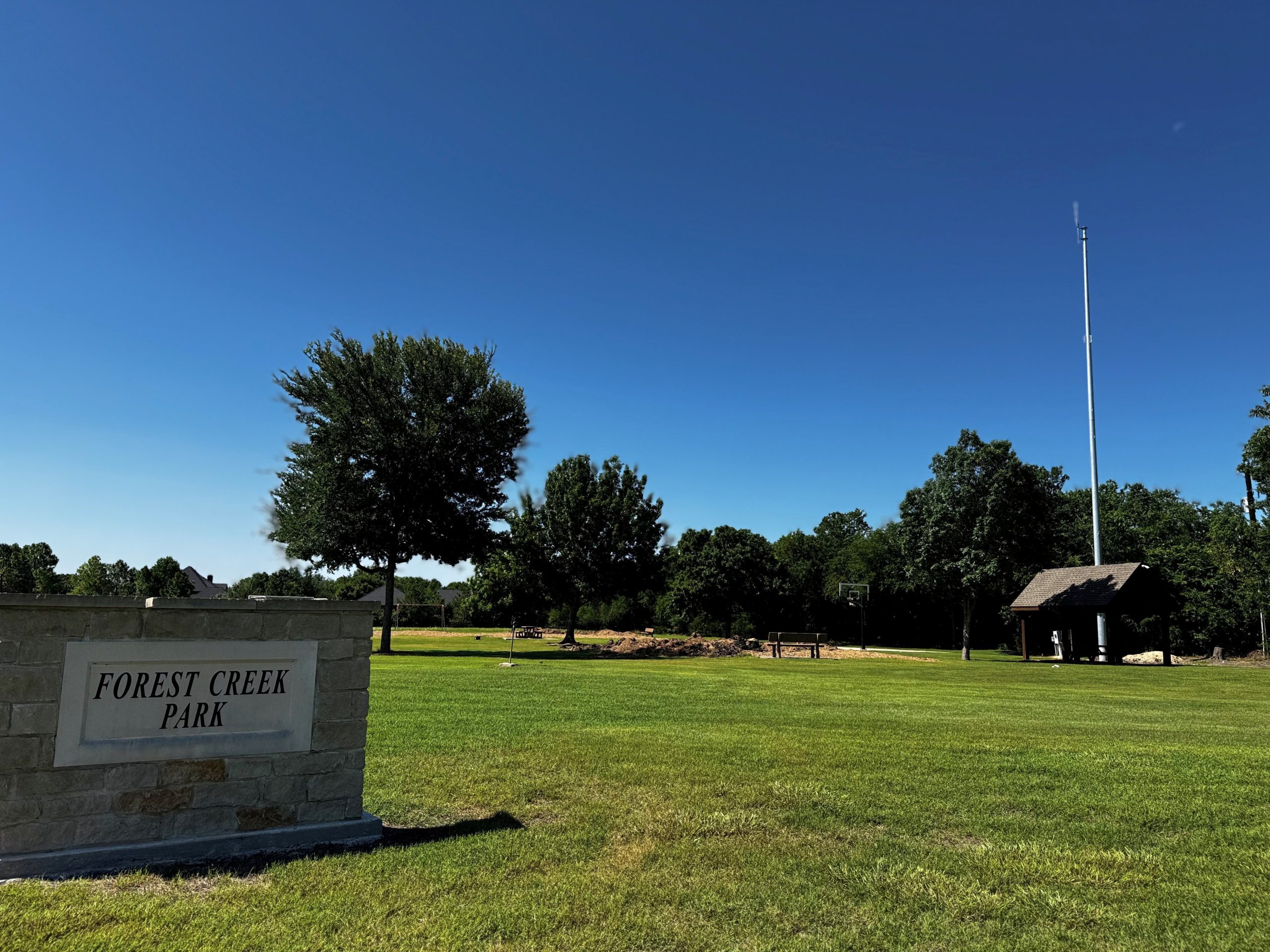 Wide-angle view of Forest Creek Park in Lucas, Texas, featuring a grassy field under a clear blue sky. A stone sign reading "Forest Creek Park" is in the foreground. In the background, there are trees, a small pavilion, a tall utility pole, and visible construction equipment where a playground once stood.