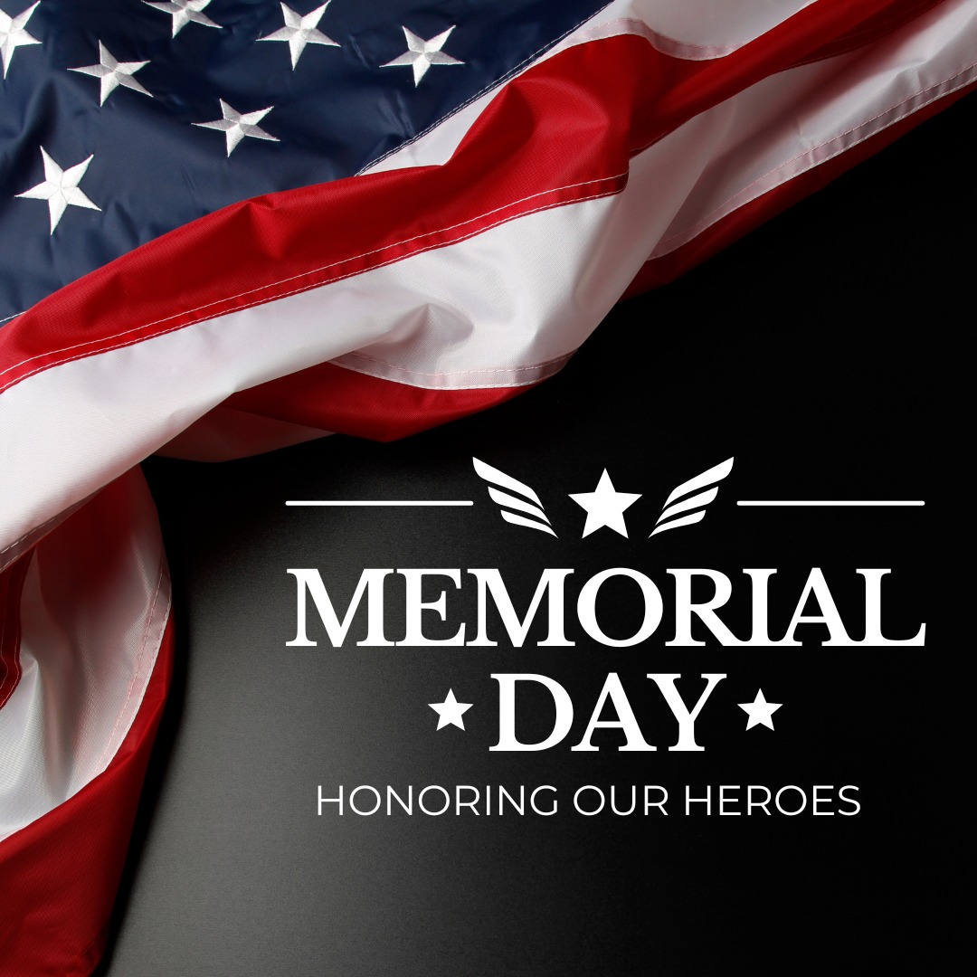 A close-up of the American flag with red, white, and blue fabric draped across a dark surface. In the foreground, white text reads "Memorial Day โ Honoring Our Heroes," with stars and wing-like graphics surrounding the words.
