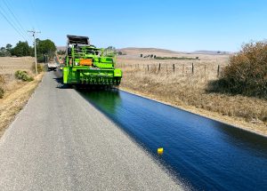 Road work being done on a rural road. A large green construction vehicle is laying fresh pavement material on the road.