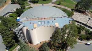 Drone photo of the top of the Marin Center's roof under construction. There is scaffolding along the walls and a construction worker fixing something on the roof.