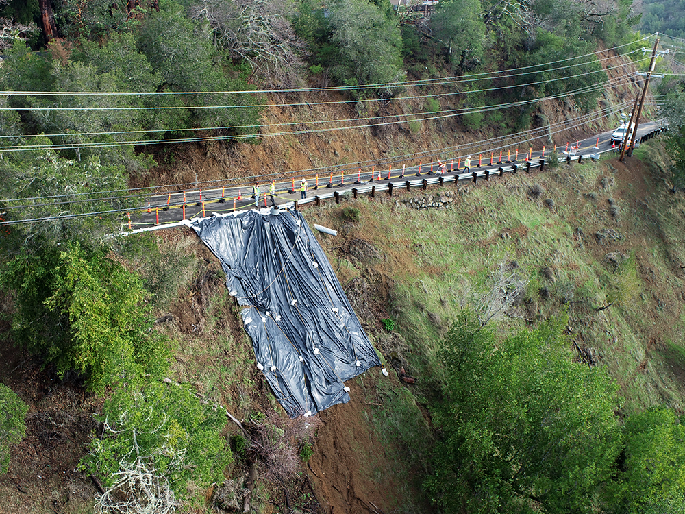 Road along a ridge with trees. The road is damaged and a black tarp has been placed to secure a landslide.