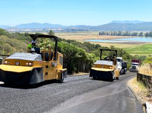 Pavement Preservation project with heavy equipment construction vehicles working on Atherton Oaks Drive in Black Point. There is a nice view of the hills and a body of water in the background behind the roadway.