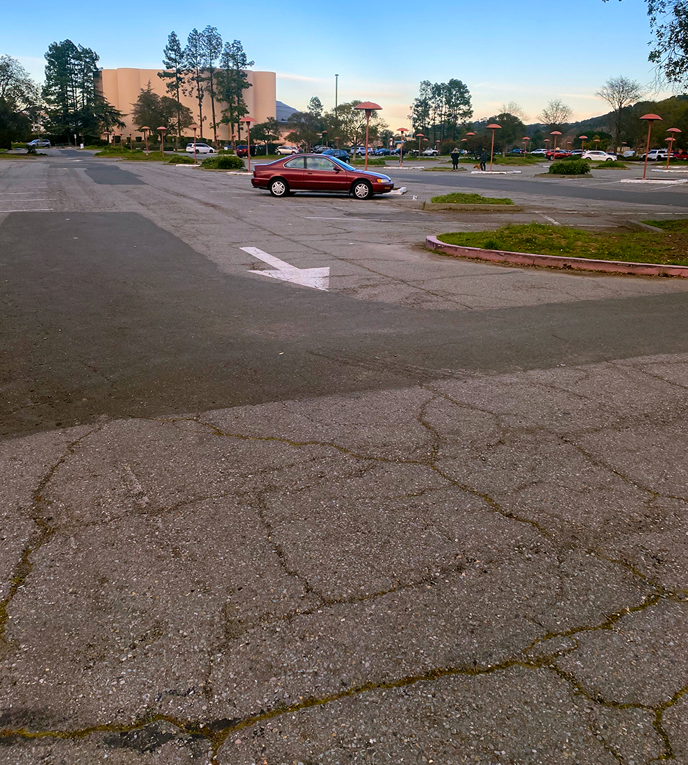 Marin Center parking lot with deteriorated pavement. The Marin Center building is visible in the background. There are a couple cars parked in the parking lot.