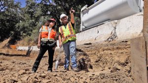 There are two construction workers at a retaining wall construction site. One of them is pointing at something off screen.