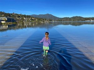 Child in rainboots stands with back to camera, walking through water atop a fully flooded bike path.