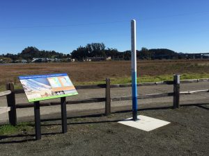 interpretive sign board and seven foot poll with banded colors stand alongside bikepath