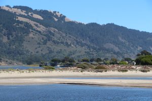 Blue lagoon waters and a vegetated sand dune are backed by steep scrub and forested ridge slopes