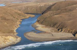 Coastal Estuary with sand bar aprtially closing river mouth draining into the Pacific Ocean. River banks are steep brown undeveloped hills.
