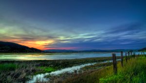 colorful sunset over a wide creek with grassy banks. Fence posts sit in the foreground, and a hill in the background.