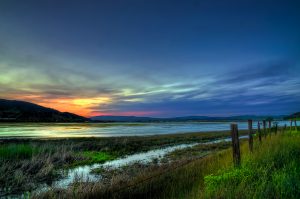 colorful sunset over a wide creek with grassy banks. Fence posts sit in the foreground, and a hill in the background.