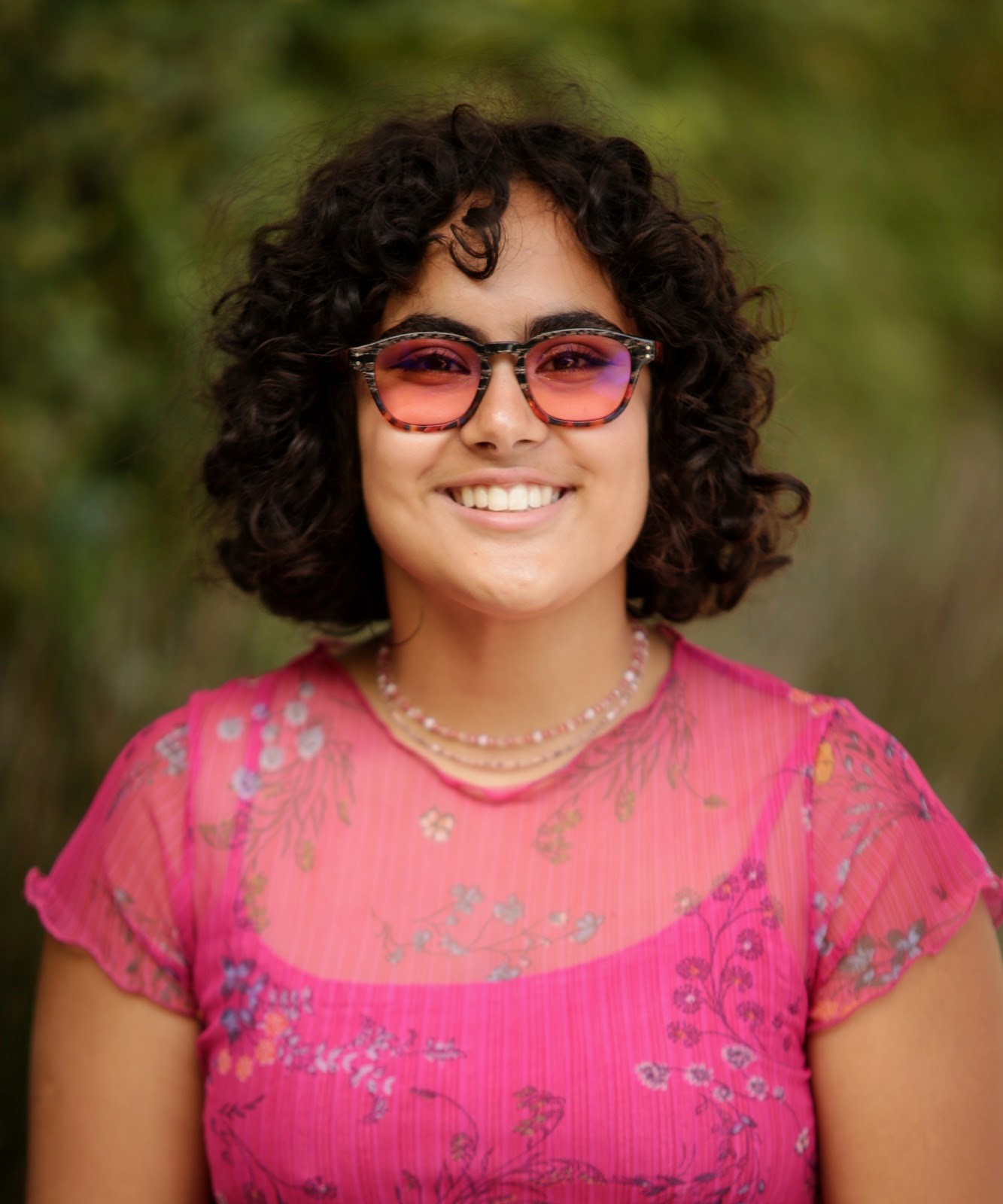 smiling young person with curly chin-length hair wearing a pink shirt and pink tinted glasses