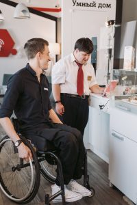 A man with down syndrome in a red tie and a man in a wheelchair in all black are looking at an item in the workplace.