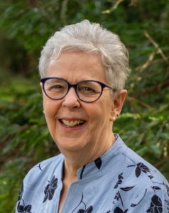 A woman with short white hair and dark glasses smiles in front of of greenery outside