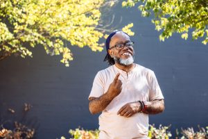 A Deaf Black man smiles while signing. The man has glasses, a beard, and septum piercing, and stands outdoors in front of a black wall with foliage