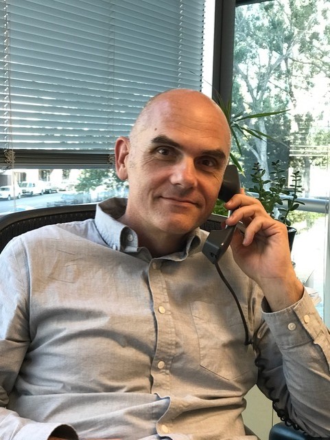 A man in a gray button up shirt sitting in an office holding a phone to his ear