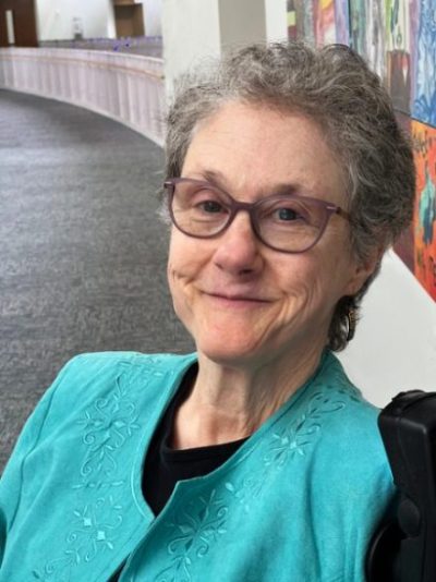 A woman with short "salt and pepper" wavy hair and glasses smiles in hallway with art on the wall