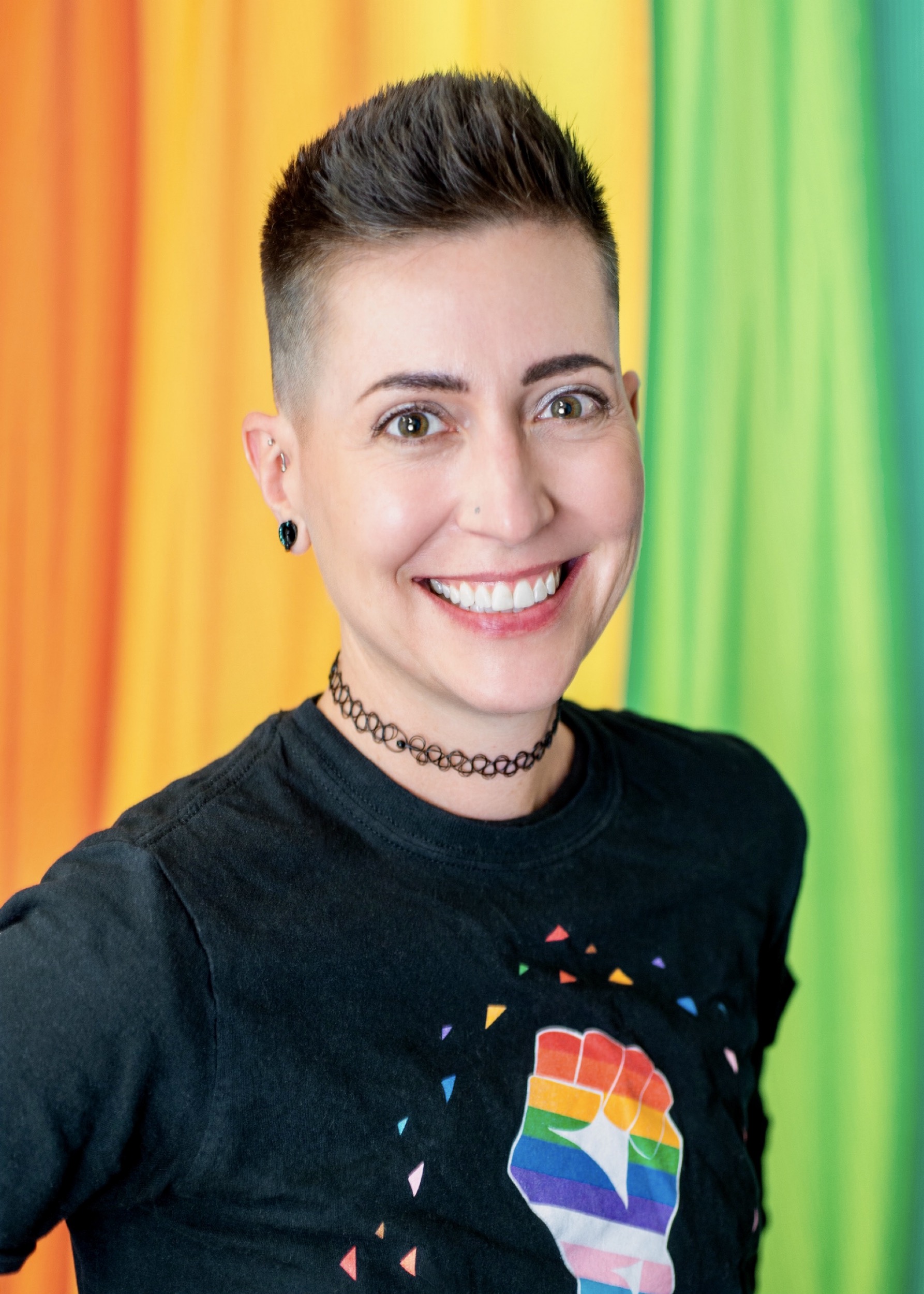 A person with short dark hair wearing a shirt with a rainbow fist smiles in front of a colorful background